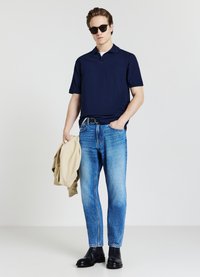 Uomo con occhiali da sole neri, maglietta polo blu navy, jeans blu e stivaletti neri, mentre tiene una giacca beige nella mano sinistra contro uno sfondo bianco.