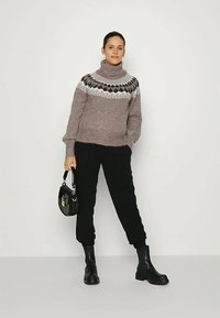 Mulher vestindo um suéter de gola alta padrão taupe, calças pretas, botas pretas até o tornozelo, segurando uma bolsa preta, de pé contra um fundo branco.