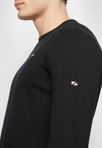 Stranski pogled na osebo, ki nosi črno majico z dolgimi rokavi in majhno belo, rdečo in modro logo na zgornjem delu roke.