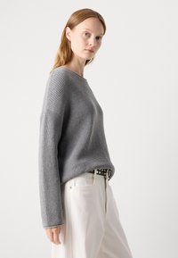 Vrouw met lichtbruin haar, gekleed in een grijze gebreide trui, witte broek en een zwarte gesp met studs, staande tegen een effen lichte achtergrond.
