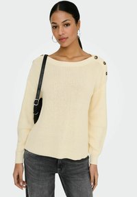 Femme portant un pull en maille crème avec des boutons sur les épaules, un jean gris foncé, des boucles d'oreilles créoles et un sac à bandoulière noir.