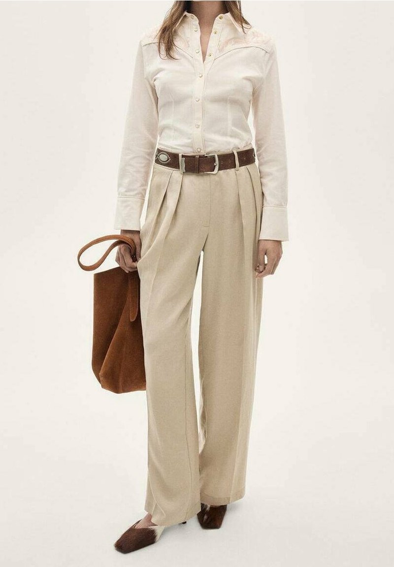 Femme portant une chemise crème à boutons, un pantalon beige taille haute à jambes larges avec ceinture marron, tenant un sac fourre-tout marron, et des chaussures marron à bout pointu.