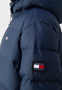 Marinblå puffjacka med kviltat mönster, med en svart lapp med varumärket "TOMMY JEANS" på ärmen.