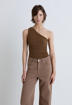 Giovane donna dai capelli castani corti, indossa un top marrone monospalla e pantaloni a vita alta marrone chiaro, in piedi davanti a uno sfondo semplice.