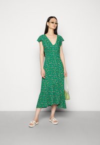 Vestido midi floral verde com mangas curtas e esvoaçantes, decote em V e bainha em camadas. Combinado com sandálias brancas e uma pequena mala verde.