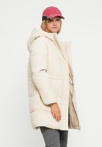 Beige dunjakke med hætte, lynlås og horisontalt quiltet design. Kombineret med en rød og sort cap, vist mod en hvid baggrund.