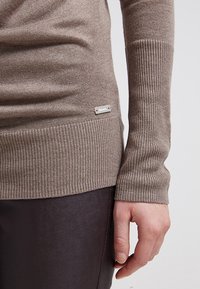 Bruine geribbelde trui met lange mouwen, voorzien van een zilveren merklabel. Het materiaal lijkt zacht en is aansluitend bij de taille, gecombineerd met donkere broeken.