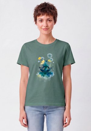 Woman with short brown hair wears green T-shirt with blue watercolor silhouette meditating amid yellow flowers and dandelions, paired with light jeans.