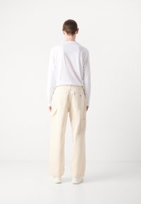 Pantalon large beige clair en tissu résistant, avec deux poches latérales, une poche arrière, et un petit détail d'étiquette sur la ceinture.