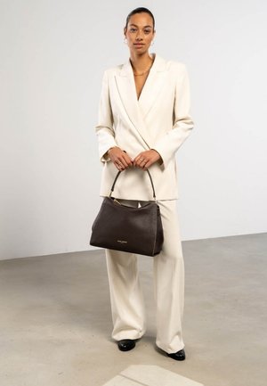 Frau in einem cremefarbenen Hosenanzug, die eine große dunkelbraune Ledertasche hält, steht in einem minimalistischen Innenraum mit neutralem Hintergrund.