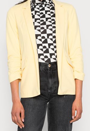 Person iført en lysegul blazer over en sort-hvid ternet rullekrave med hjerter og sorte højtaljede jeans, synlig fra skuldre til hofter.