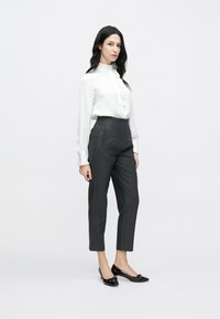 Balta blūze ar volāniem, kombinēta ar tumši pelēnām, augsta vidukļa piegriezuma biksēm un spīguļojošām melnām kurpēm ar dekoratīvām lokām, uz vienkārša fona.