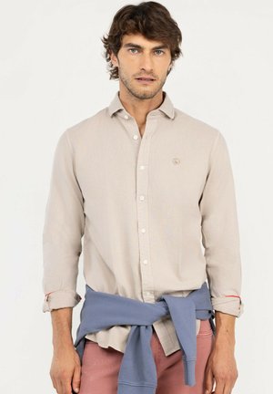 Hombre con las mangas remangadas lleva una camisa beige abotonada, sudadera azul atada alrededor de la cintura y pantalones rosas, de pie contra fondo blanco.