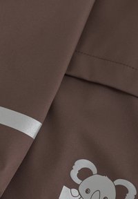 Brun tyg med en mjuk textur, prydd med en silvergrafik av en koala och en horisontell silvrig rand. Slät material och sömmar synliga.