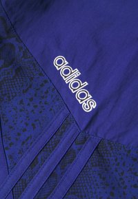 Sezione giacca Adidas in vivace tessuto viola con un motivo nero testurizzato, accentuata dal logo Adidas ricamato in bianco.