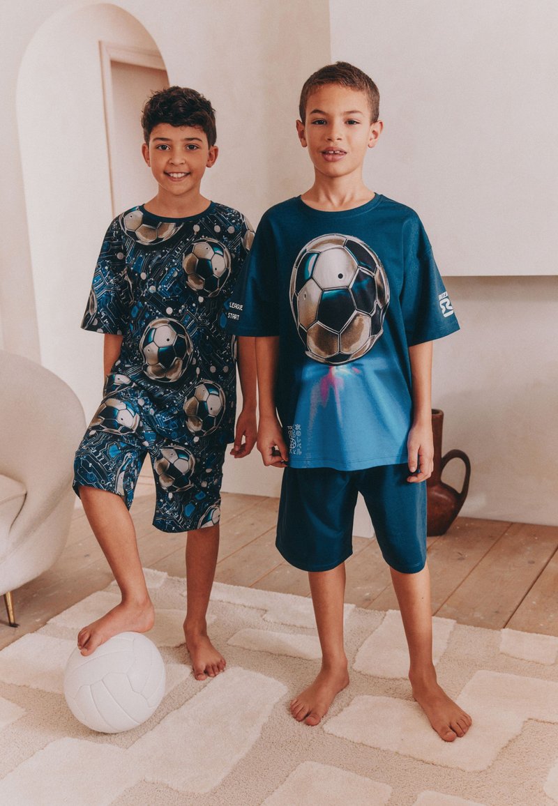 To barefødte drenge står indendørs på et mønstret tæppe iført matchende fodboldtema t-shirts og shorts, den ene med en hvid fodbold ved sin fod.