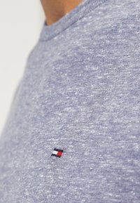 Ljust blått bomullströja med en strukturerad yta. Har en liten broderad logotyp i svart, rött och vitt på vänster bröst.