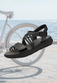 Czarny sandał wykonany z teksturowanego materiału, z plecionymi paskami, płaską podeszwą i regulowanym zapięciem. Lekka konstrukcja z minimalnym wykończeniem.