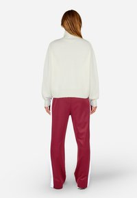 Kremowy sweter z golfem o luźnym kroju i prążkowanymi mankietami, zestawiony z bordowymi spodniami dresowymi z białymi paskami po bokach i prostymi nogawkami.