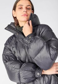 Giacca puffer grigia con colletto alto, trapuntatura a rombi, tasche con zip e dettagli a bottone a pressione. Il tessuto ha una finitura liscia e lucida.