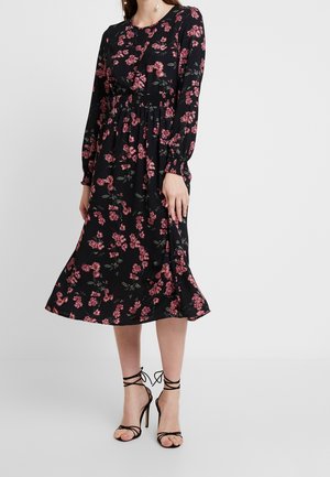 Femme portant une robe midi noire à manches longues avec un imprimé floral rose et des sandales noires à talons avec des brides à la cheville.
