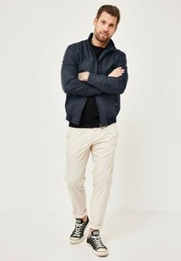 Navy bomber jacket with a zip front, black t-shirt, cream pants, and black sneakers. Model stands with arms crossed against a plain background.