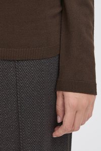 Gros plan sur la main d'une personne reposant près de l'ourlet d'un pull en maille marron et d'un pantalon gris à motif chevron.