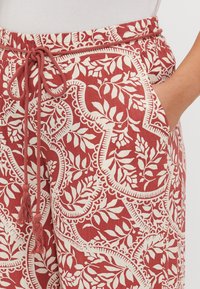 Pantaloni cu model roșu și crem, cu talie elastică cu șnur, buzunare și un design floral cu detalii intricate. Textură de țesătură moale.