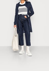 Femme portant un trench-coat bleu marine, un pull rayé en tricot, un jean large foncé, des bottines blanches épaisses, tenant un sac fourre-tout beige dans la main droite.
