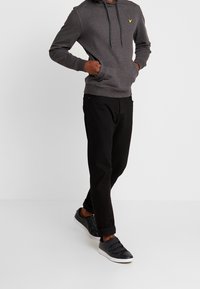 Szary sweter z kapturem z przednimi kieszeniami i sznurkiem, noszony z czarnymi spodniami i czarnymi butami sportowymi na rzepy.