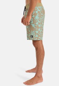 Pantalones cortos de surf en color beige arenoso con patrones de hojas tropicales en turquesa. Incluye un parche de logo negro y una cintura con cordón.