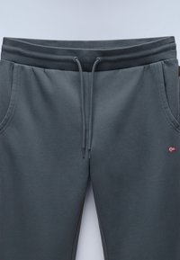 Donkergrijze joggingbroek met een elastische tailleband, een trekkoordsluiting, twee zijzakken en een klein vlaggenlabel aan de rechterkant.