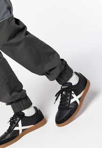 Sneakers in pelle nera con accenti bianchi, suola in gomma color beige, abbinati a joggers grigi con vita elastica, caratterizzati da polsini arricciati e una finitura liscia.