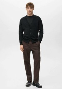 Czarny kardigan z guzikami, noszony na czarnej koszuli, w połączeniu z brązowymi spodniami i czarnymi butami. Materiał wydaje się gładki i miękki.