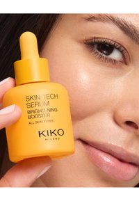 Ljusgul dropperflaska märkt "KIKO Skin Tech Serum Brightening Booster" som hålls nära en persons ansiktshud. Slät, klar textur.