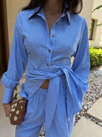Camisa riscada azul claro com aplicações de pérolas, frente abotoada, mangas longas, atada na cintura. Combinada com calças de padrão correspondente e uma mala texturizada.