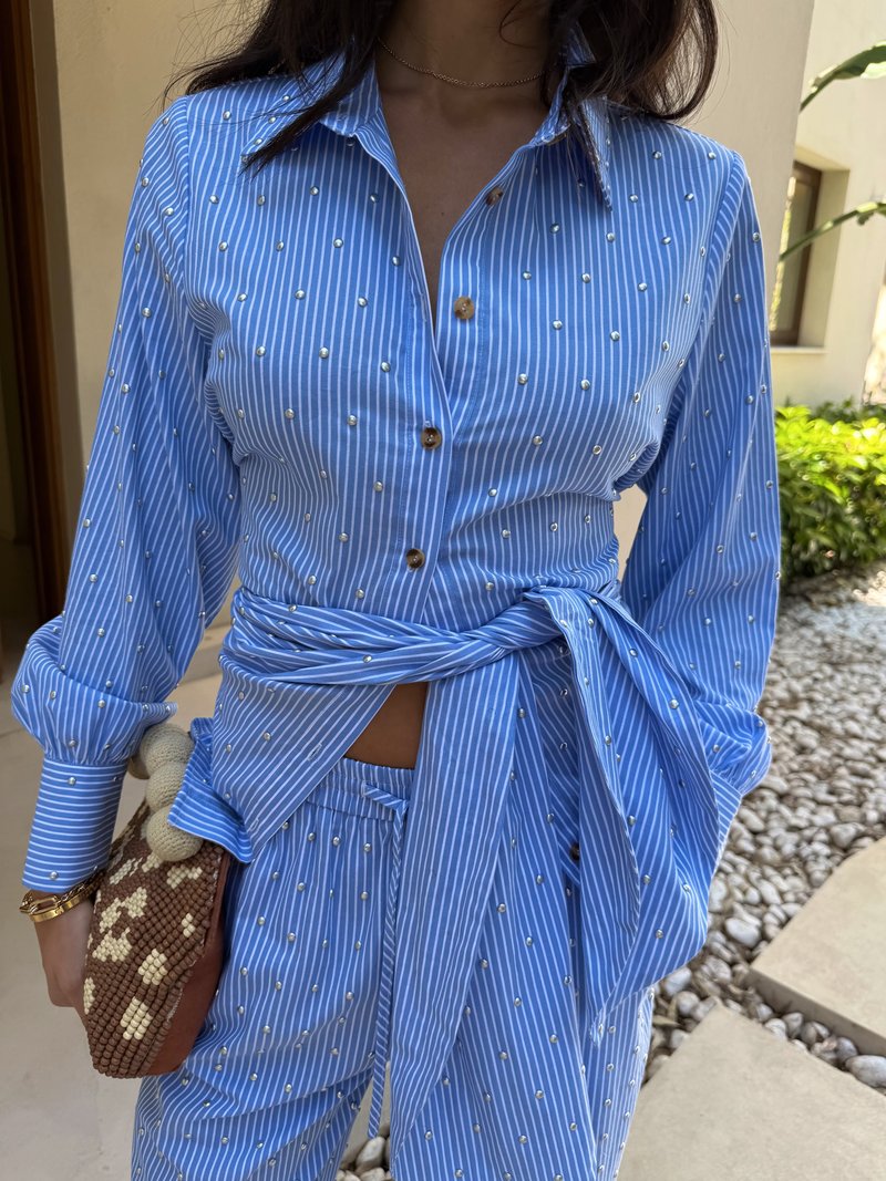 Camisa riscada azul claro com aplicações de pérolas, frente abotoada, mangas longas, atada na cintura. Combinada com calças de padrão correspondente e uma mala texturizada.