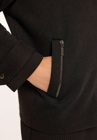 Czarna kurtka z teksturowanej tkaniny, z zamkniętą kieszenią i detalami na mankietach. Zamek ma metaliczny wykończenie z małą metką marki.