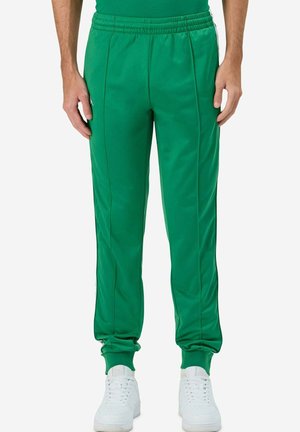 Pantaloni tuta in tessuto verde e liscio con vita elastica, strisce laterali e polsini aderenti, abbinati a scarpe da ginnastica bianche.