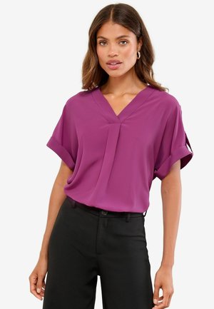 Blusa a maniche corte con scollo a V in tessuto viola brillante. Presenta risvolti alle maniche e una lieve plissettatura sul davanti. Indossata con pantaloni neri.