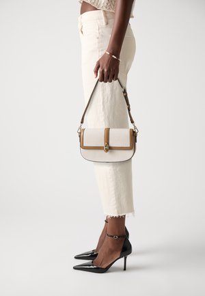 Crossbody bag with a textured cream fabric, brown trim, and gold hardware. Black pointed heels and light-colored cropped jeans complete the look.