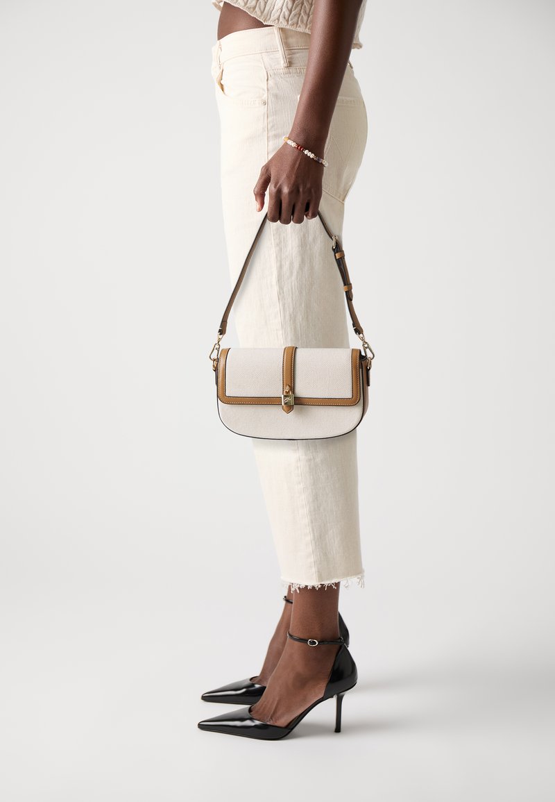 Crossbody bag with a textured cream fabric, brown trim, and gold hardware. Black pointed heels and light-colored cropped jeans complete the look.