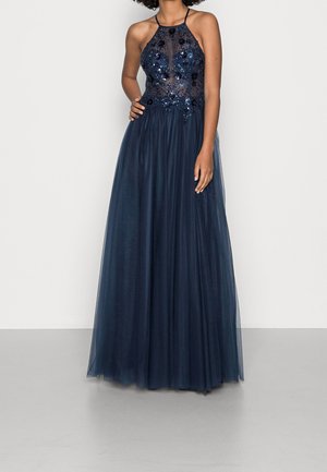 Femme portant une robe de soirée bleu marine sans manches avec un corsage transparent et orné, et une jupe en tulle fluide, debout avec une main sur la taille.