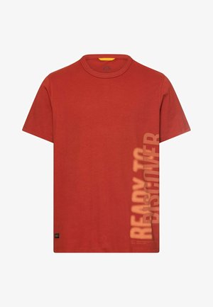 T-shirt à manches courtes couleur rouille avec col rond et texte vertical délavé "PRÊT À DÉCOUVRIR" sur le côté droit près de l'ourlet.