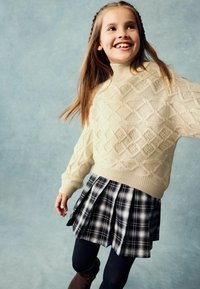 Pull en tricot de couleur crème avec des motifs de diamants texturés, associé à une jupe plissée à carreaux noir et blanc et des leggings noirs. Des bottes marron viennent compléter la tenue.