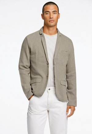 Lichtbeige blazer van een gestructureerde stof, met opstaande revers, twee voorzakken en bruine knopen, gecombineerd met een grijs shirt en witte broek.