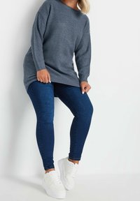 Suéter de punto gris con un corte holgado y mangas largas, combinado con jeans ajustados de color azul oscuro y zapatillas blancas. Textura suave y estilo casual.