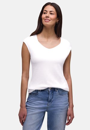 Jeune femme portant un haut blanc sans manches et un jean bleu, regardant légèrement vers sa droite avec un fond neutre.