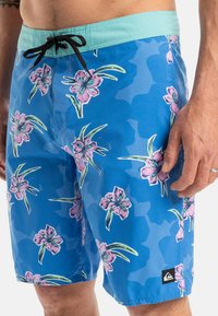 Blå blommiga badbyxor med en turkos midja, med ett camouflage-mönster och svart dragsnöre. Signaturlogotyp på den nedre sidan.