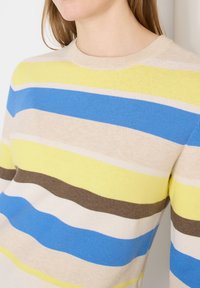 Femme portant un pull à col rond avec des rayures horizontales jaunes, bleues, beiges, blanches et marron.